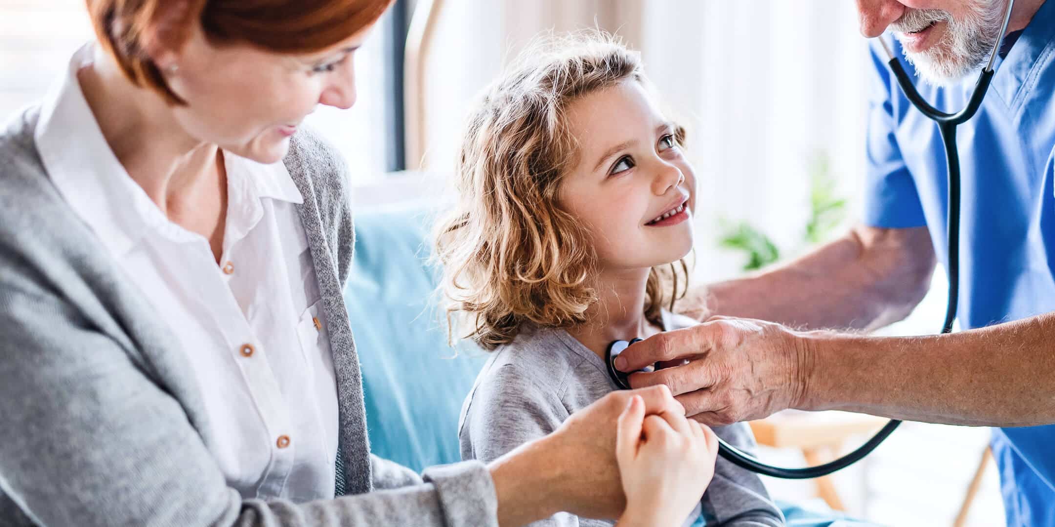 Ung pige, der sidder på sin mors skød under en lægekonsultation og smiler
