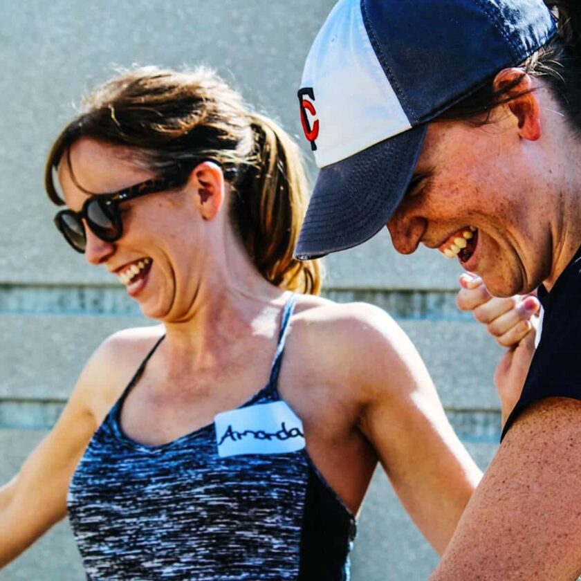 To kvinder griner sammen udendørs. Den ene bærer solbriller, den anden kasket. Navneskiltet siger "Amanda".