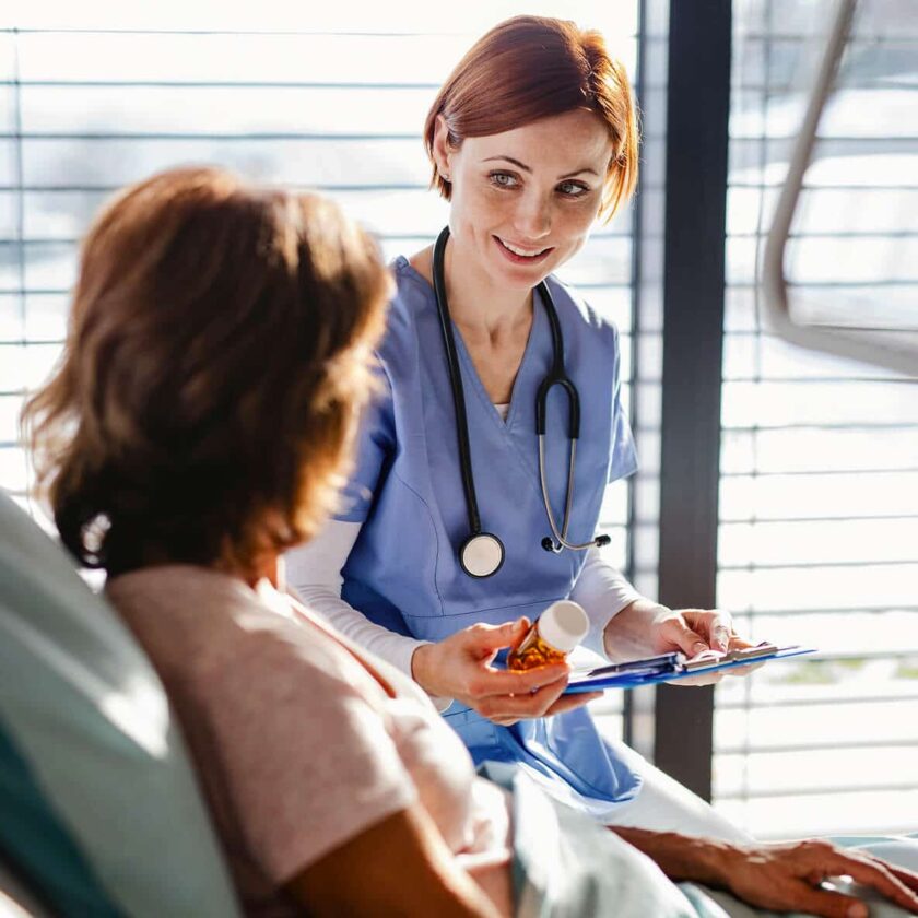 Sygeplejerske taler med patient i seng, holder medicinglas og clipboard, begge smiler.
