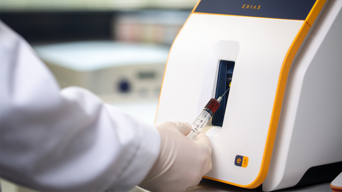 Handske iført hånd indsætter en blodprøve-sprøjte i en medicinsk analyser-maskine mærket "EXIAS" til testning.