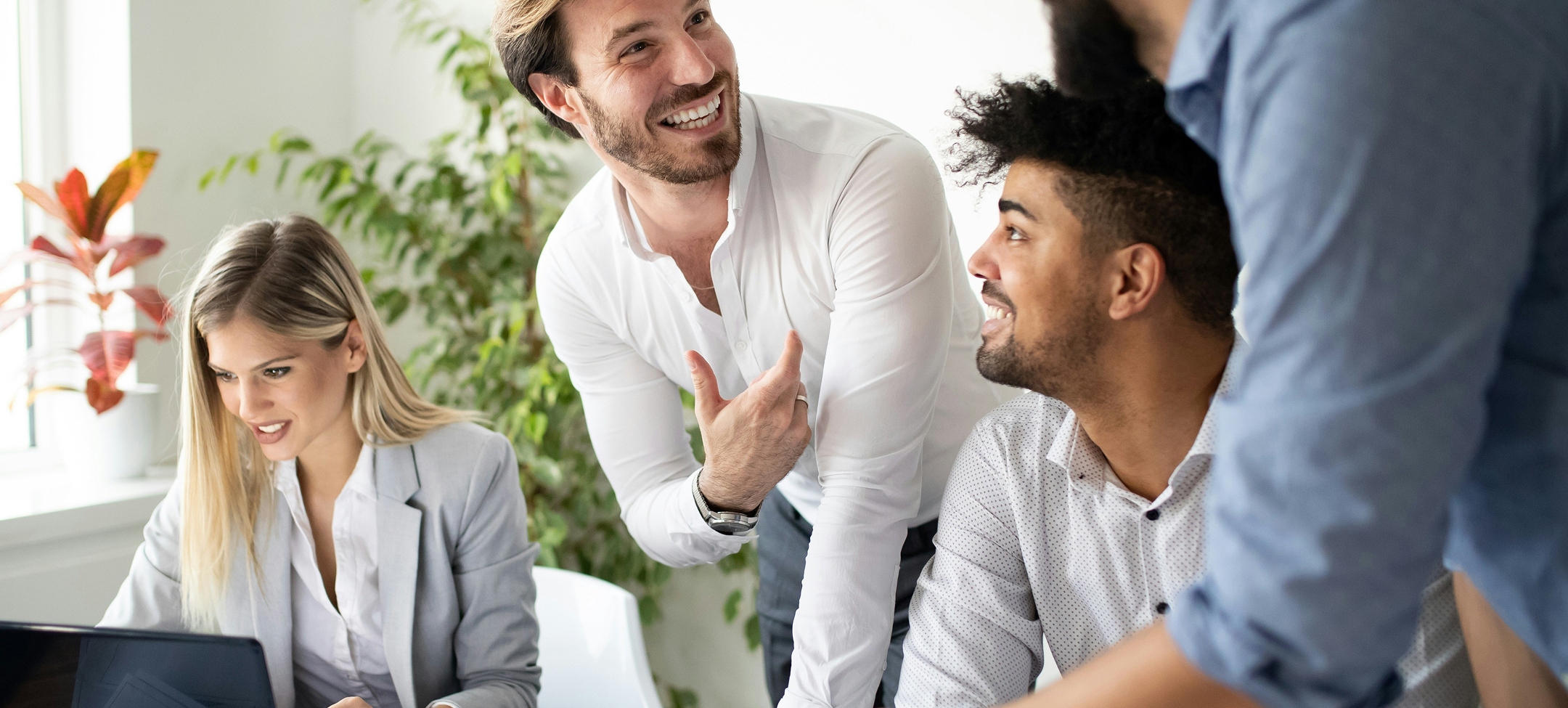 Fire kolleger har en samtale og smiler sammen på et kontor med grønne planter og en bærbar computer.