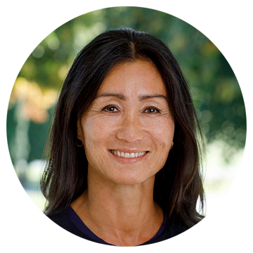 Kvinde med langt mørkt hår smiler foran en sløret baggrund med grønne træer.