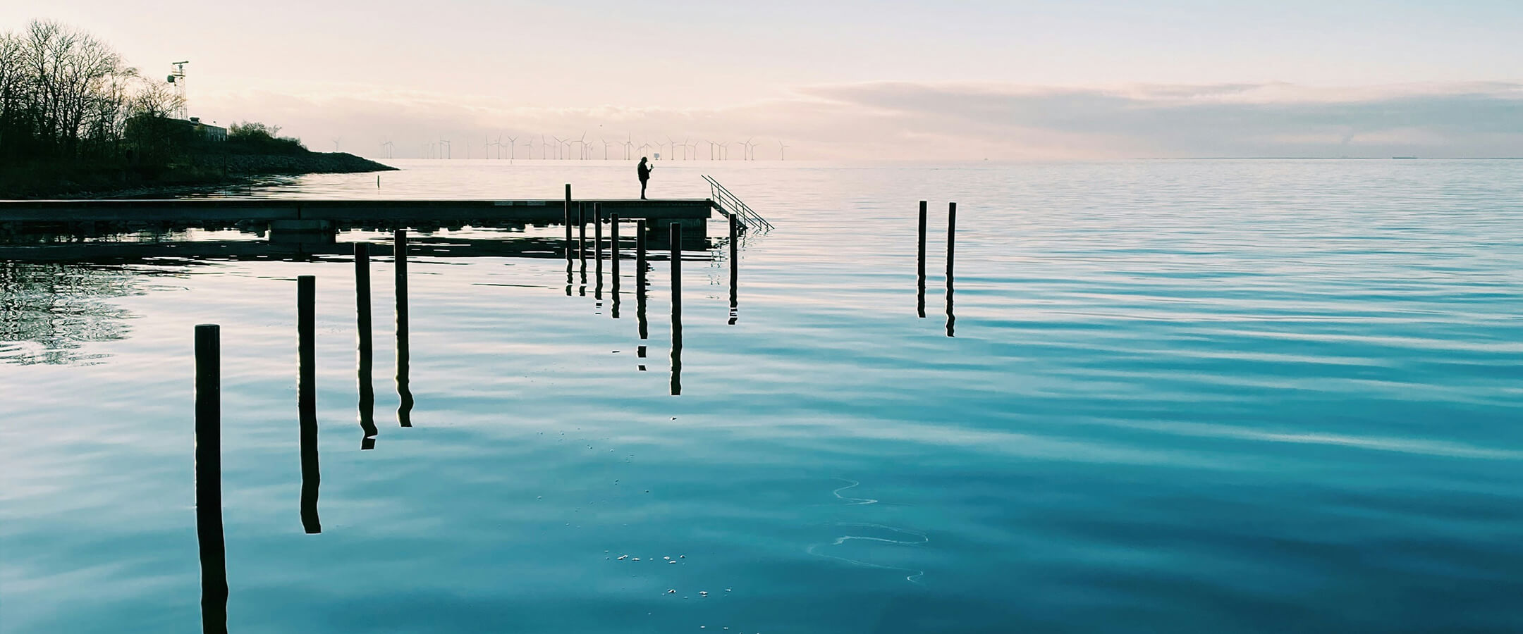 Blå, rolig sø med badebro, en person står for enden. Vindmøller ses i horisonten under en lys himmel.