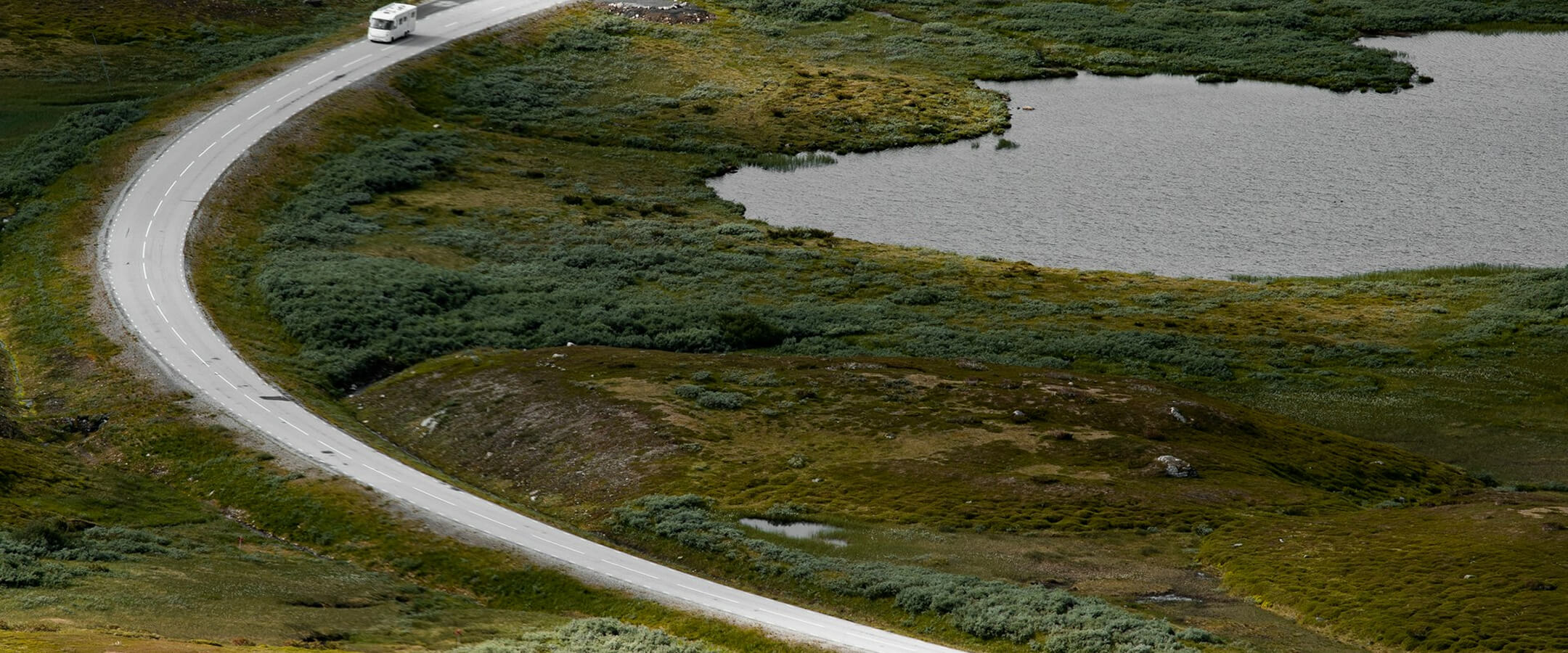 Blandet landskab med snoet vej, autocamper og sø omgivet af grøn bevoksning.