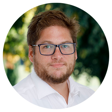 Mand med brunt hår, briller og skæg iført hvid skjorte, sløret grøn baggrund.