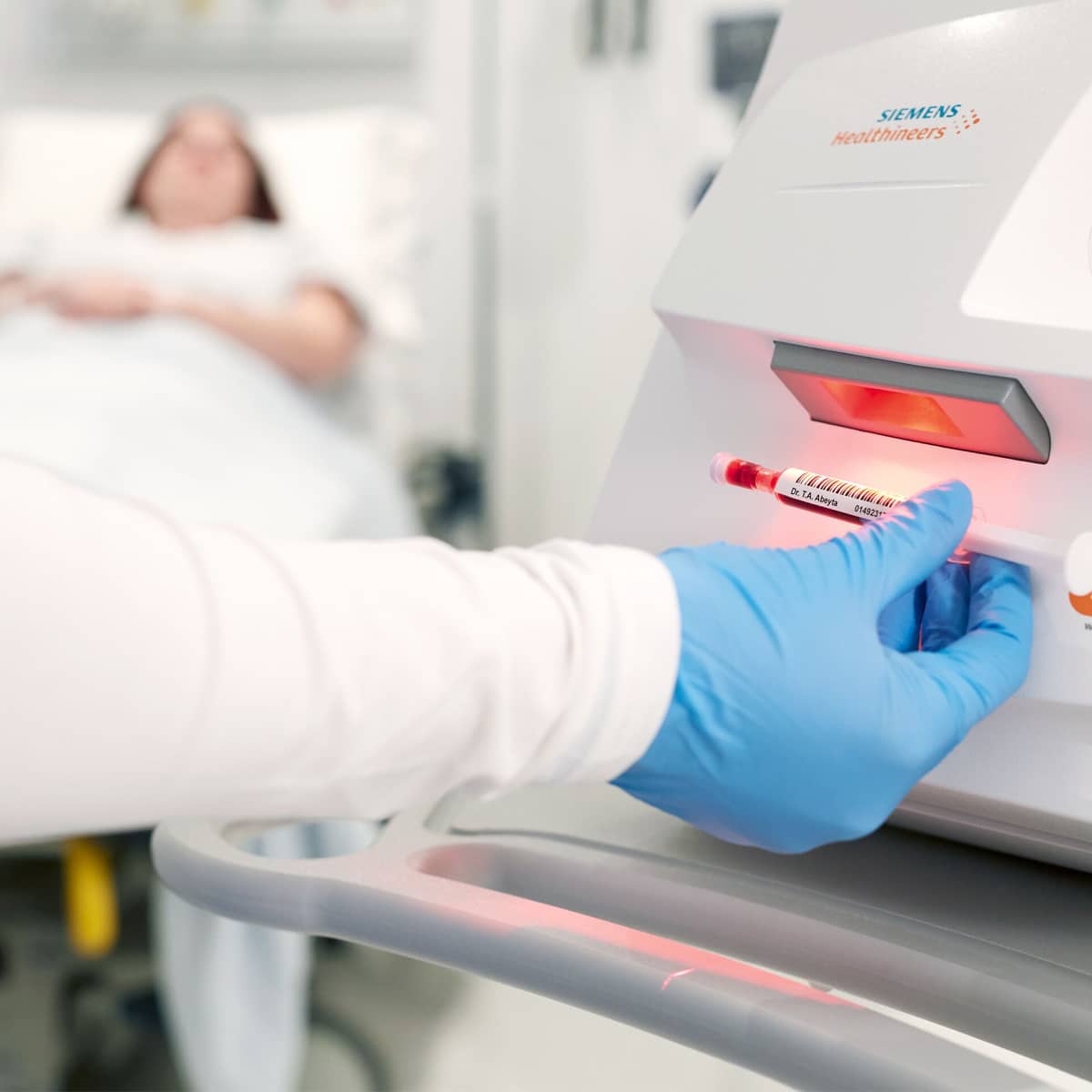 Hånd med blå handske indsætter blodprøve i Siemens maskine, patient ligger i baggrunden på hospitalsseng.