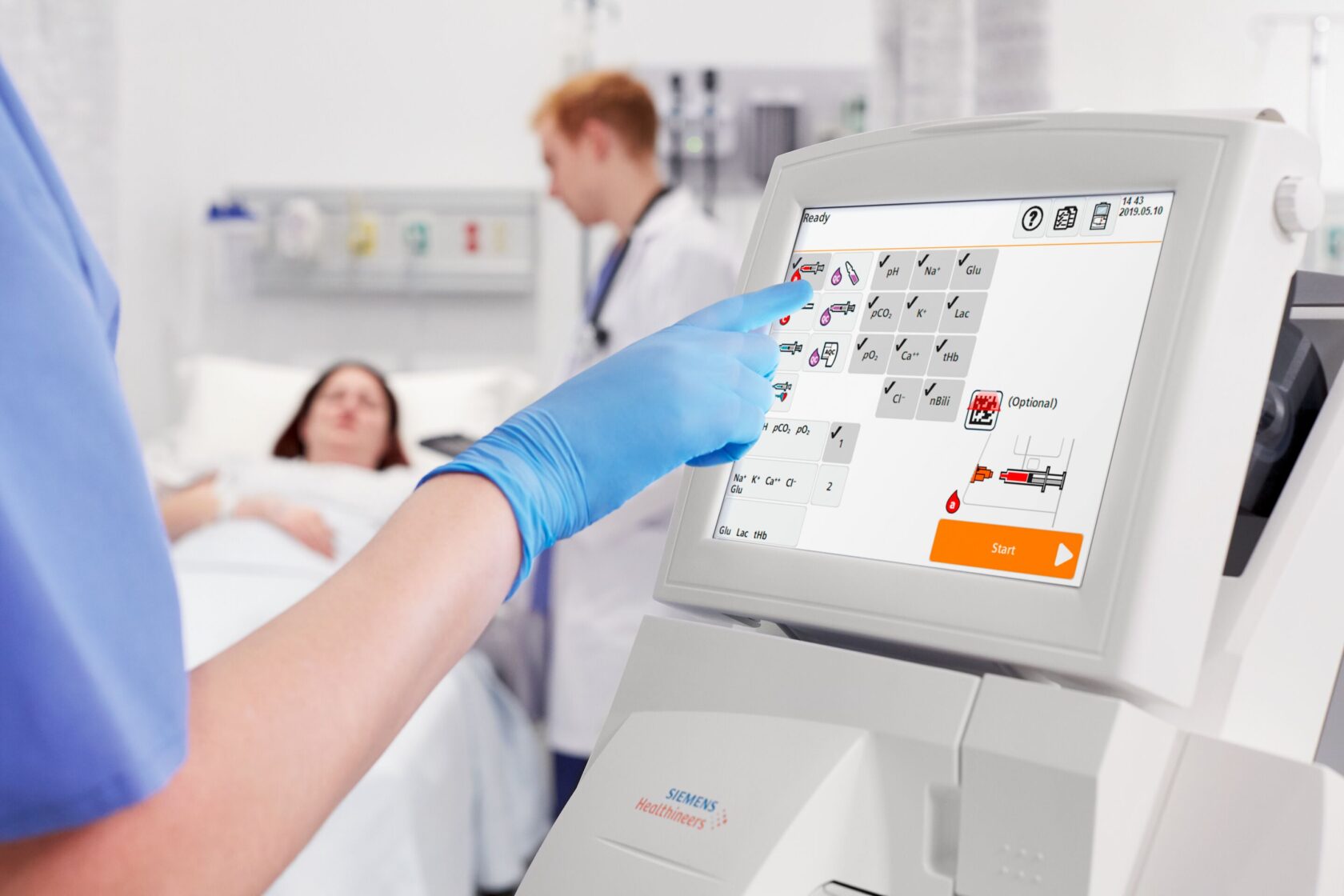 En sundhedsarbejder bruger en Siemens medical touchskærm, patient og læge ses i baggrunden på sygehusværelse.