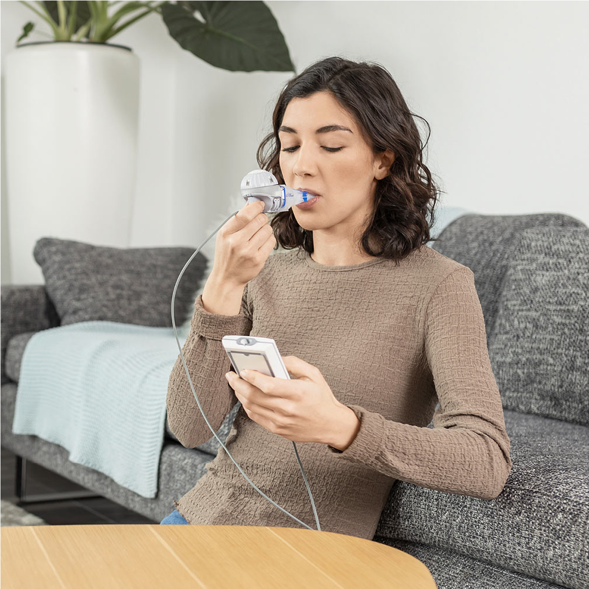 Kvinde bruger en medicinsk åndedrætsenhed, mens hun sidder på en sofa og holder en kontrolenhed.