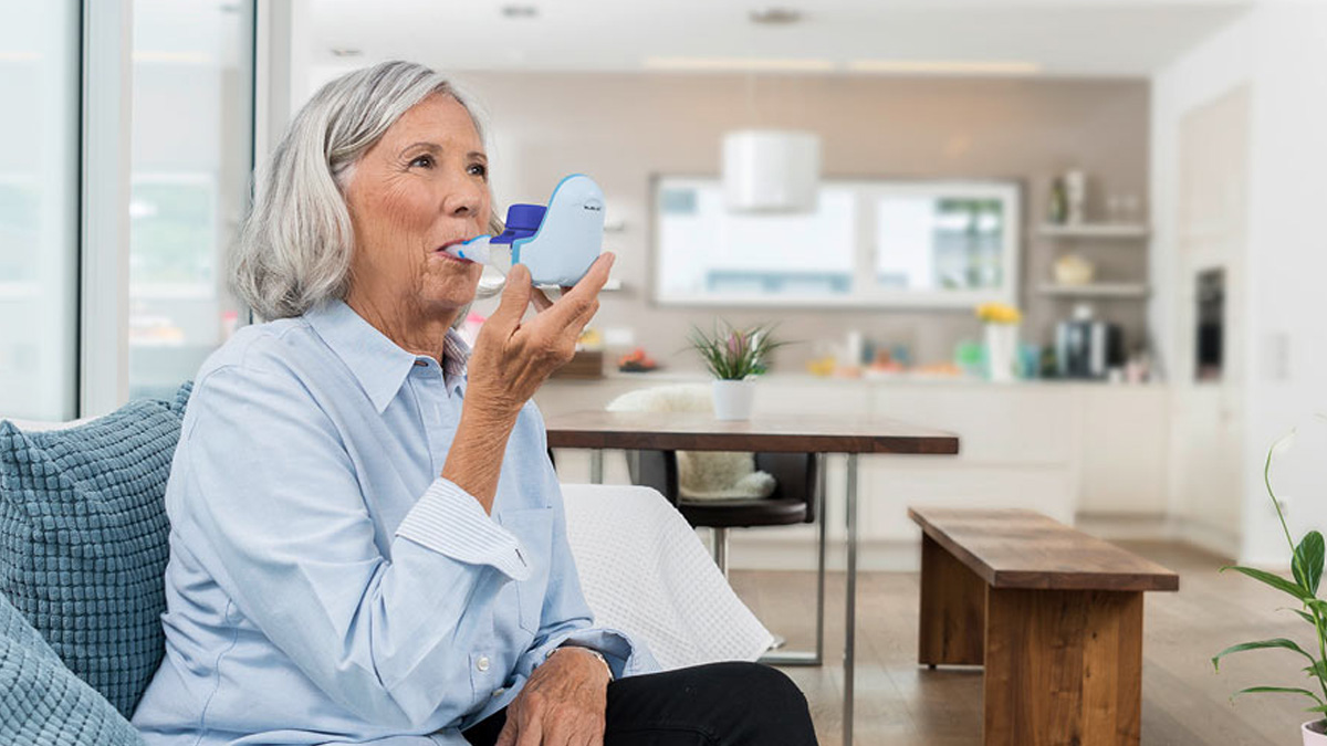 Ældre kvinde siddende i en sofa og bruger en blå respiratorisk inhalator i et lyst, moderne køkken.