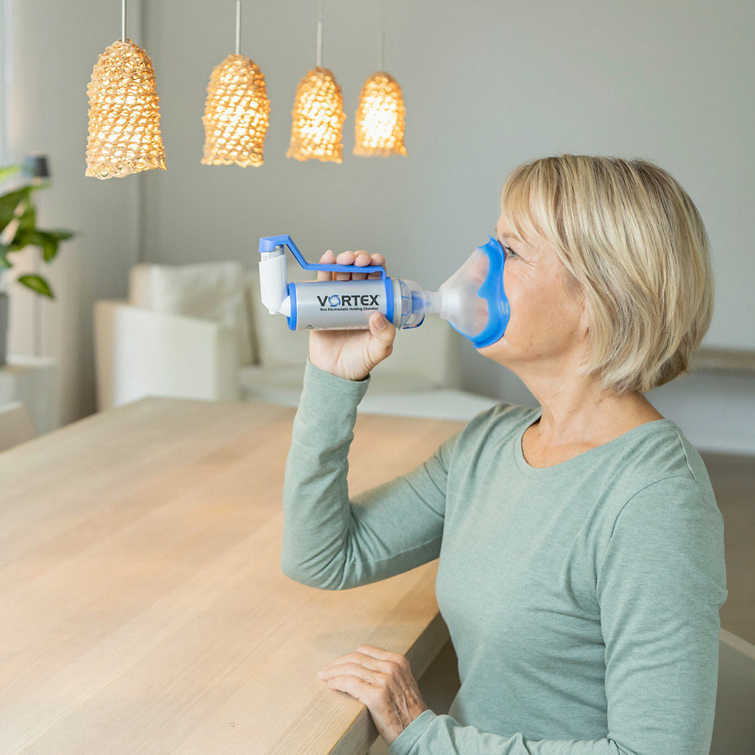 Kvinde, der bruger en inhalator med spacer ved et bord i et veloplyst, moderne rum med hængelamper.