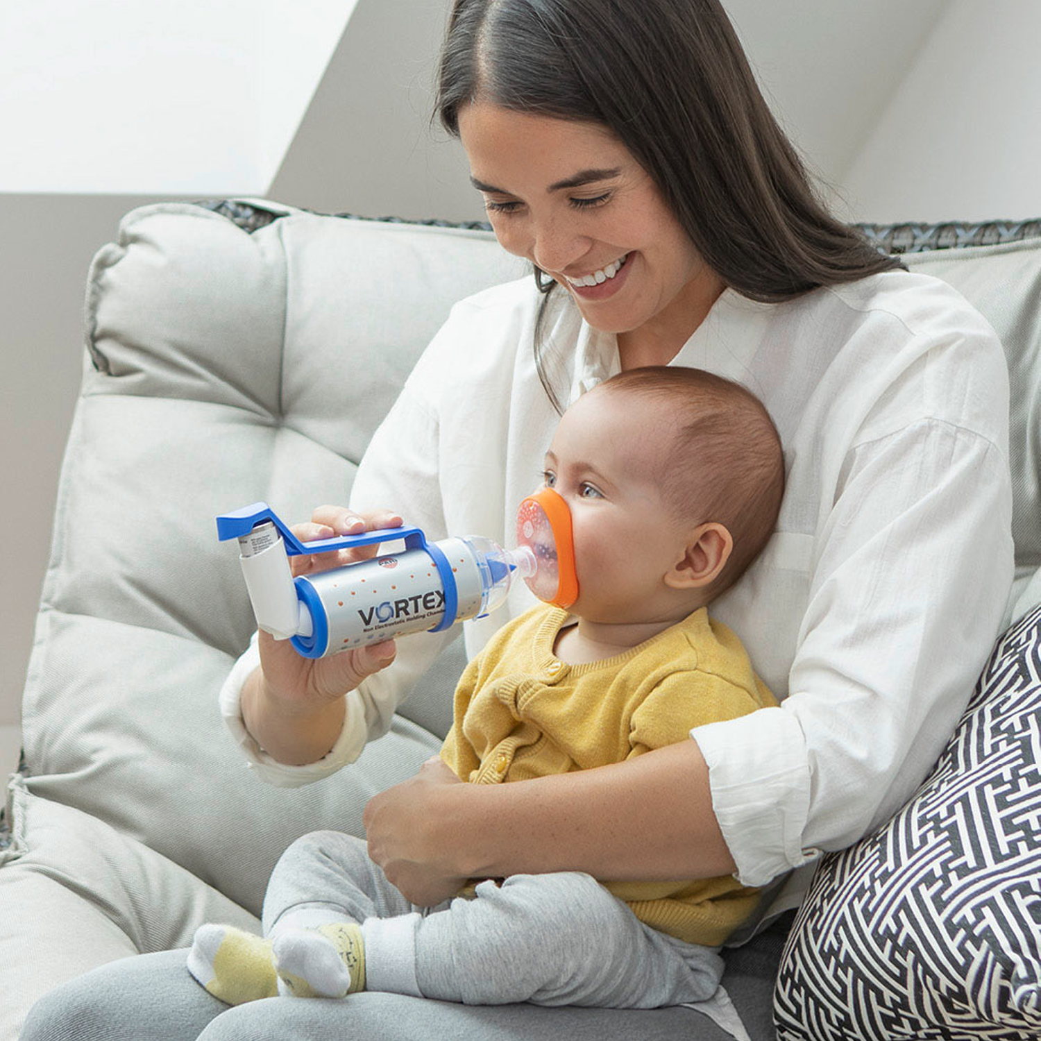 Kvinde smilende, mens hun giver en baby en inhalator med maske, siddende på en sofa.