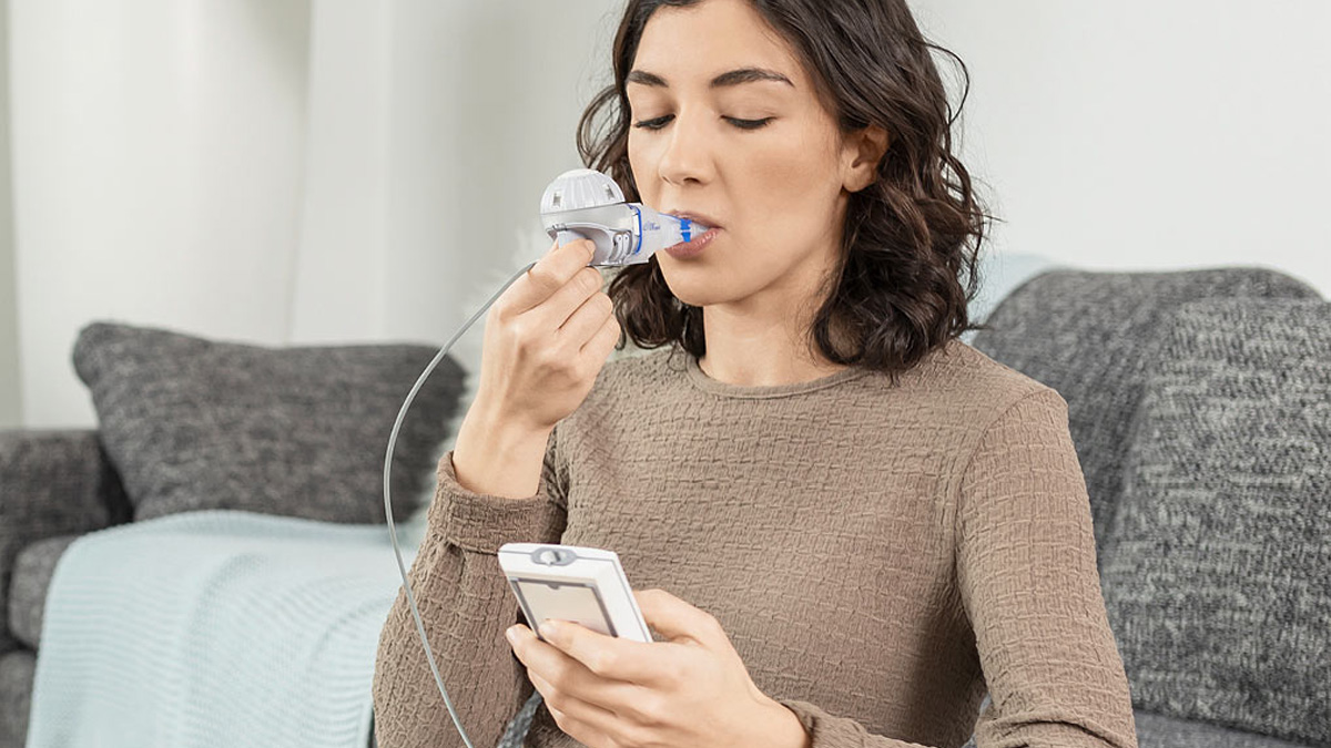 Kvinde bruger en håndholdt respirationsenhed, mens hun sidder i en sofa og holder en tilsluttet mobilenhed.