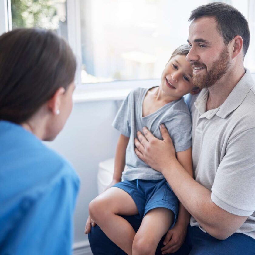 Læge, der konsulterer en ung dreng, som sidder på sin fars skød på et hospital