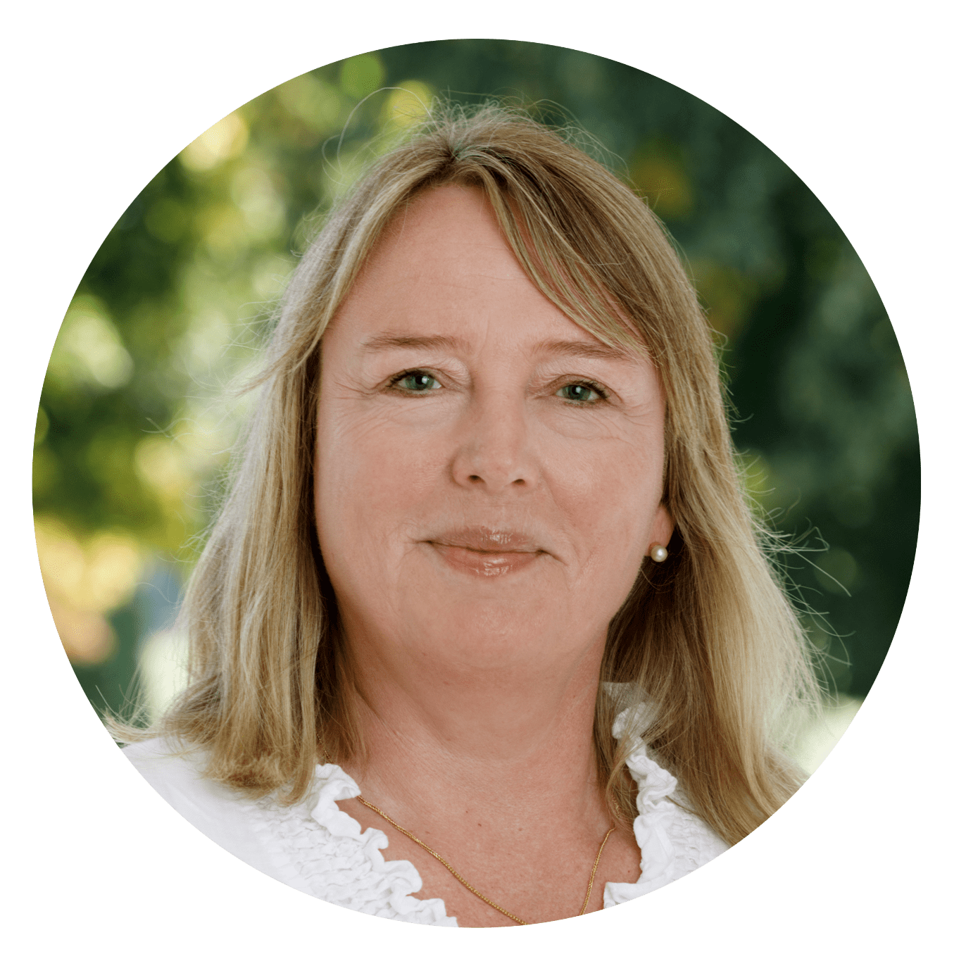 Kvinde med lyst hår smiler, iført hvid bluse. Udendørs baggrund med grønne, uskarpe træer.