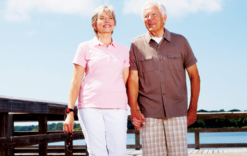 Ældre par holder i hånd og går på en træbro under en blå himmel med natur i baggrunden.