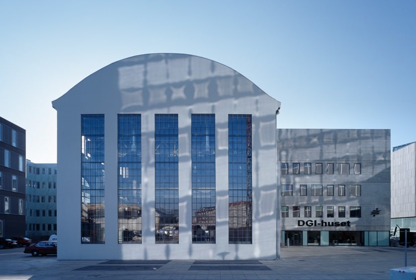 Moderne bygning med høje buede vinduer og reflekterende glas, mærket "DGI-huset" foran. Klar blå himmel ovenover.