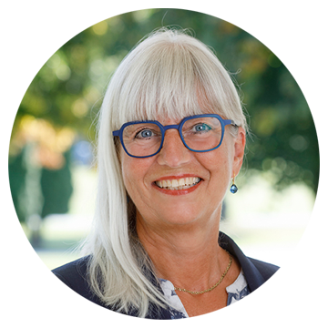 Ældre kvinde med langt, lyst hår, blå briller og blå ørering, smiler udendørs foran grøn baggrund.