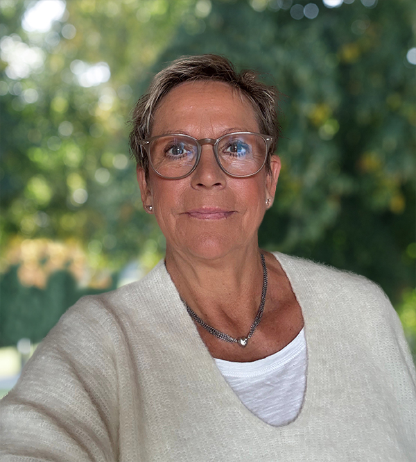 Kvinde med kort hår og briller smiler let foran grønt, uskarpt baggrund. Hun har hvid sweater og halskæde på.